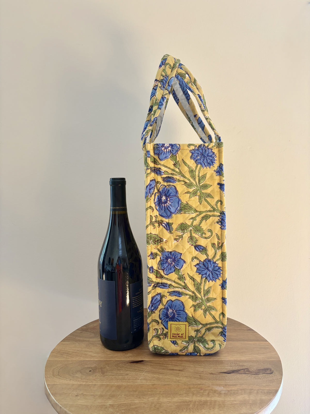 House of Mulmul - Quilted Wine Tote Bags. A yellow fabric wine bottle carrier with a blue and green floral print stands on a round wooden table next to a dark wine bottle against a plain beige wall.