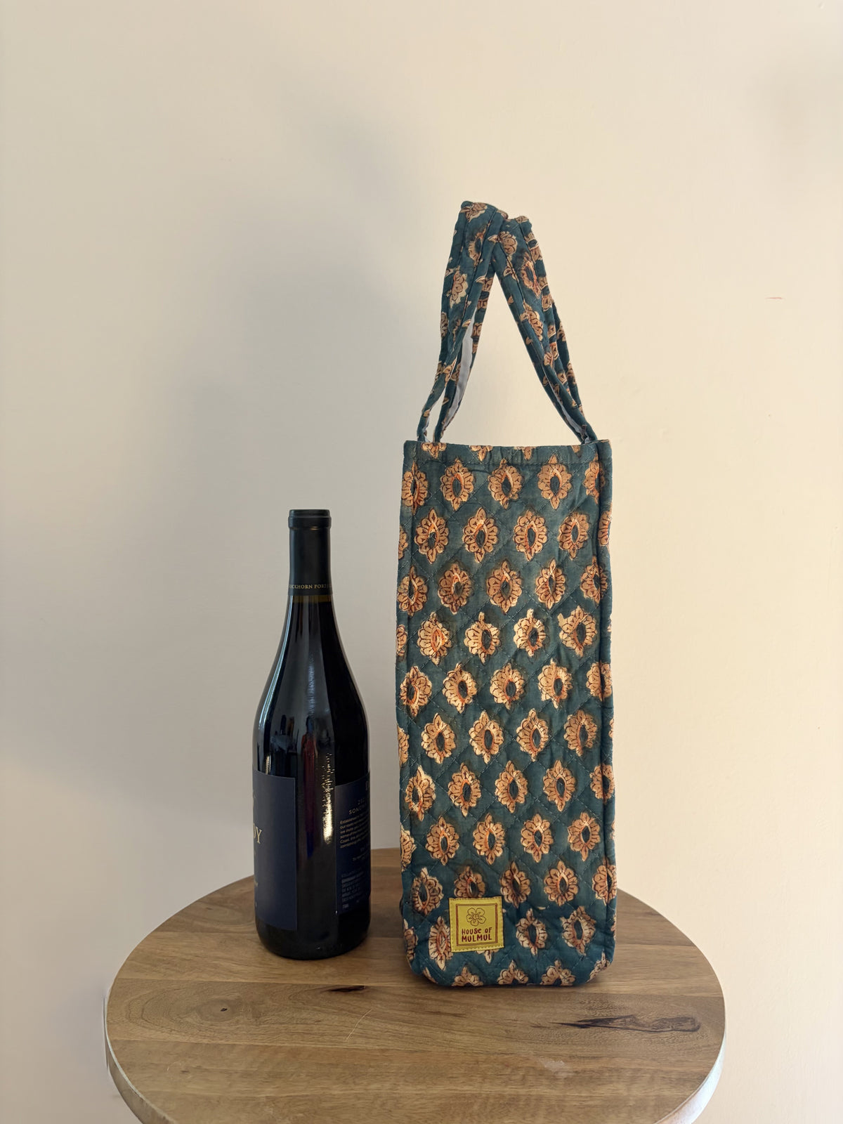 House of Mulmul - Quilted Wine Tote Bags. A patterned wine bottle carrier bag with a matching shoulder strap, displayed next to a dark wine bottle on a round wooden table against a plain beige wall.
