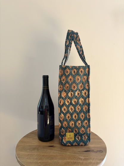 House of Mulmul - Quilted Wine Tote Bags. A patterned wine bottle carrier bag with a matching shoulder strap, displayed next to a dark wine bottle on a round wooden table against a plain beige wall.