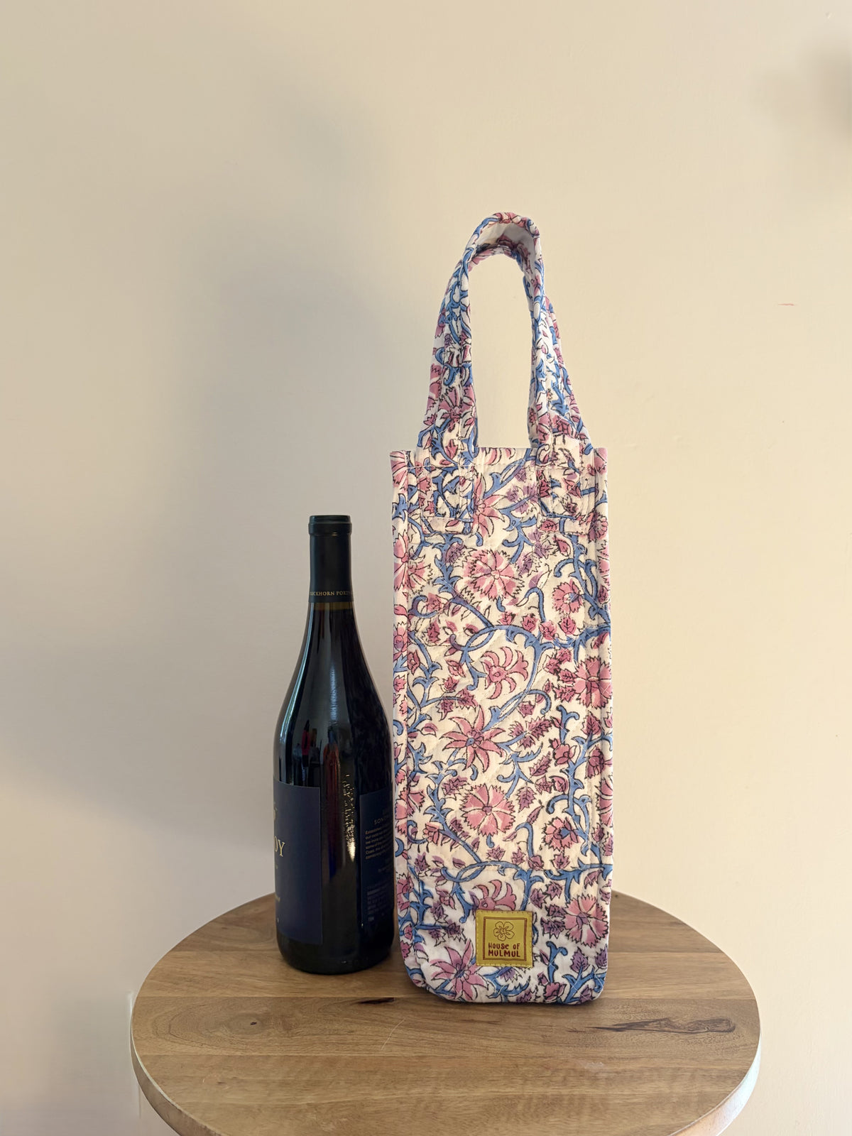 House of Mulmul - Quilted Wine Tote Bags. A floral wine bottle carrier bag with blue handles, standing on a round wooden table next to a dark wine bottle against a plain beige wall.