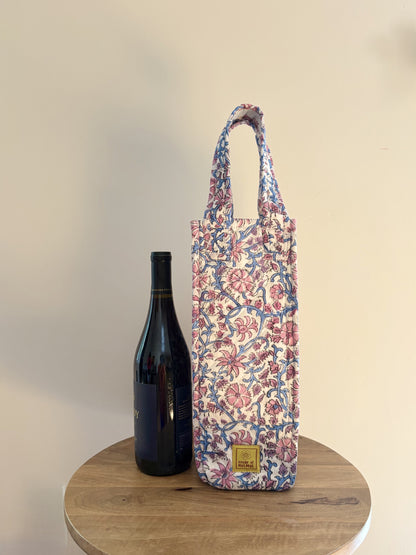 House of Mulmul - Quilted Wine Tote Bags. A floral wine bottle carrier bag with blue handles, standing on a round wooden table next to a dark wine bottle against a plain beige wall.