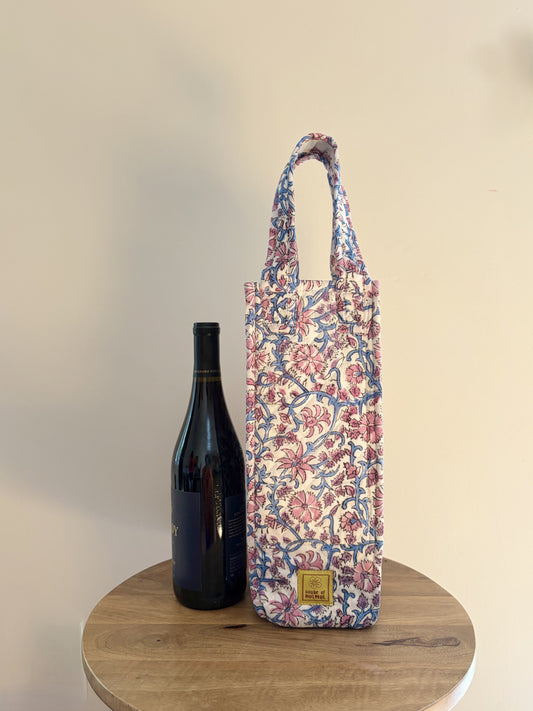 House of Mulmul - Quilted Wine Tote Bags. A floral wine bottle carrier bag with blue handles, standing on a round wooden table next to a dark wine bottle against a plain beige wall.