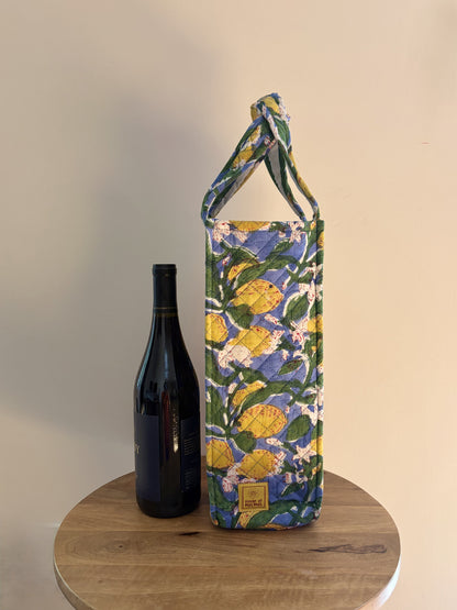 House of Mulmul - Quilted Wine Tote Bags. A fabric wine tote bag with a lemon and grapevine pattern, standing on a round wooden table next to a dark wine bottle against a plain beige wall.