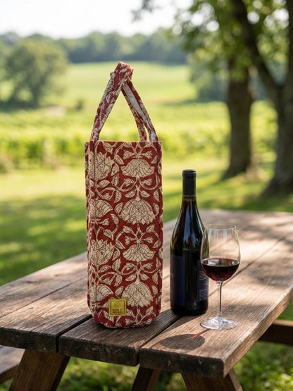 House of Mulmul - Quilted Wine Tote Bags. A red and beige patterned reusable wine tote bag stands on a wooden picnic table outdoors, next to a bottle of red wine and a filled wine glass, with a blurred green field and trees in the background.