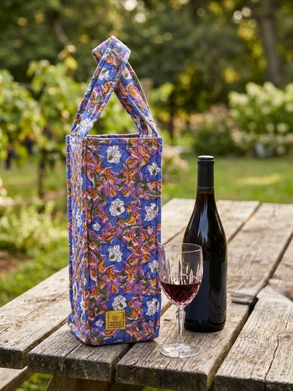 House of Mulmul - Quilted Wine Tote Bags. A floral wine bottle carrier bag with a top handle sits on a rustic wooden table outdoors, styled with a bottle of red wine and a filled wine glass. The bag features a vibrant purple, blue, orange, and white floral pattern, with a small gold brand label at the bottom.