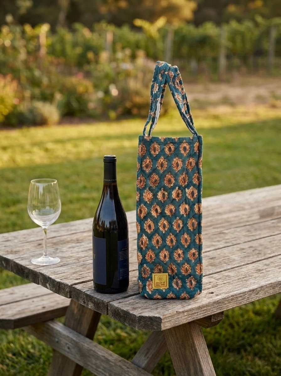 House of Mulmul - Quilted Wine Tote Bags. A patterned wine tote bag with a blue and orange geometric design stands on a rustic wooden picnic table outdoors next to a wine bottle and a wine glass, with a blurred garden background.