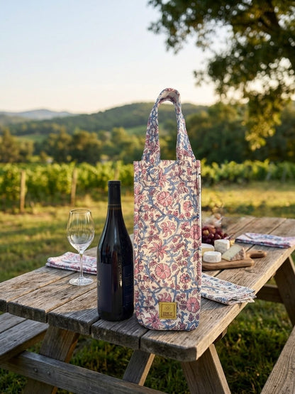 House of Mulmul - Quilted Wine Tote Bags. A floral tote bag stands on a rustic wooden picnic table outdoors, styled with a wine bottle, wine glass, cheese, grapes, and a picnic cloth. The background shows a vineyard and hills under warm sunlight.