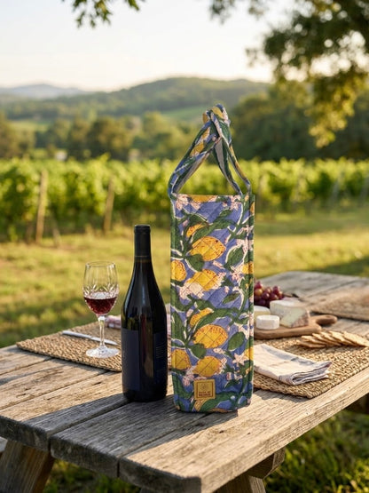 House of Mulmul - Quilted Wine Tote Bags. A patterned wine tote bag stands on a rustic wooden picnic table outdoors, styled with a bottle of red wine, a glass of red wine, cheese, crackers, and grapes. The background shows a blurred vineyard landscape under warm sunlight.