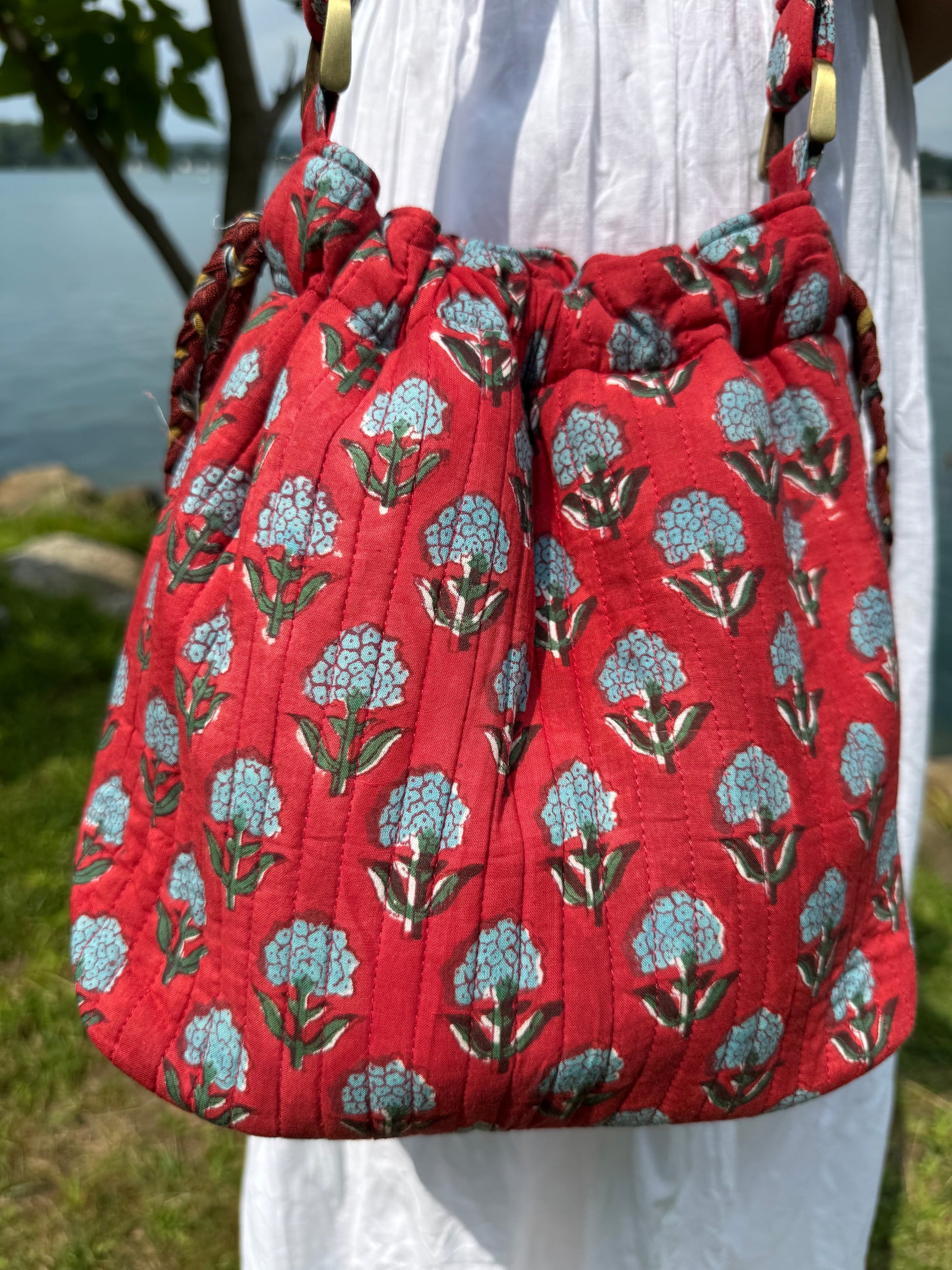 House of Mulmul - Garden Wayfarer Bucket Bag, Cotton bag, Crossbody sling bag. Red drawstring bag with floral pattern held by a person outdoors.
