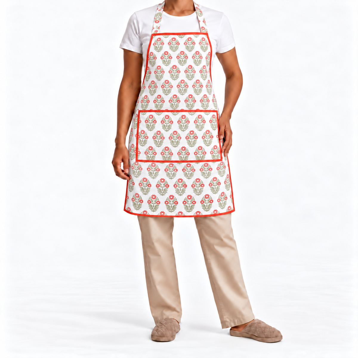 House of Mulmul - Blockprint Cotton Apron. A person wearing a white apron with a repeating coral-red and green floral block print pattern, styled over a white t-shirt and beige trousers against a plain white studio background.