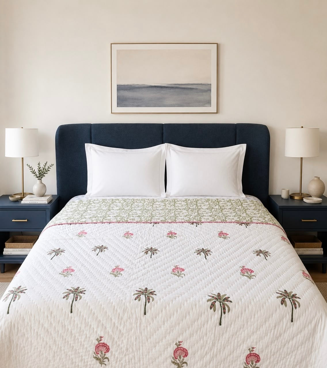 House of Mulmul - Blockprint Cotton Quilt Reversible. A neatly made bed in a styled bedroom, featuring a white quilted coverlet with a palm tree and pink floral pattern, a green patterned band, and a dark navy blue upholstered headboard. Two matching nightstands with lamps sit on either side, and a framed abstract print hangs on the wall above.
