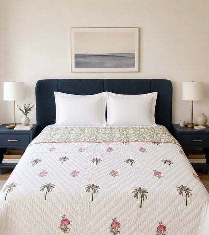 House of Mulmul - Blockprint Cotton Quilt Reversible. A neatly made bed in a styled bedroom, featuring a white quilted coverlet with a palm tree and pink floral pattern, a green patterned band, and a dark navy blue upholstered headboard. Two matching nightstands with lamps sit on either side, and a framed abstract print hangs on the wall above.