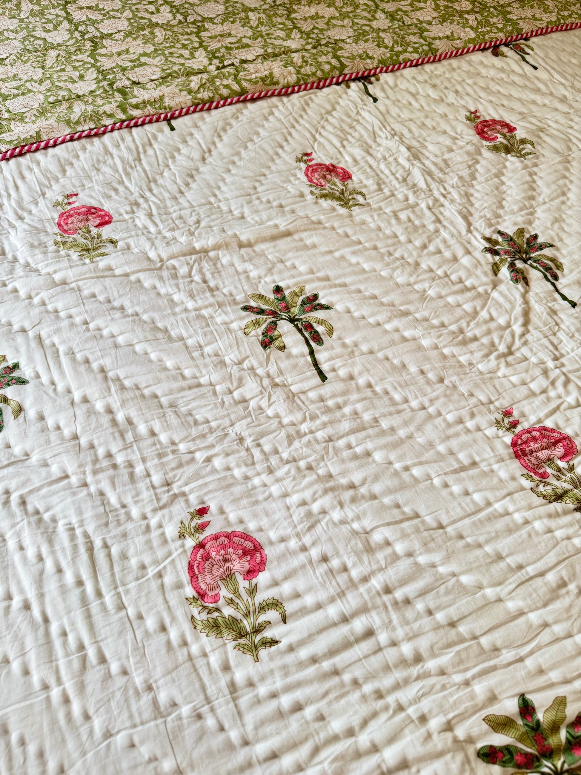 House of Mulmul - Blockprint Cotton Quilt Reversible. A close-up of a quilted white cotton blanket featuring embroidered pink flowers and green leaves, with a red and white twisted rope border along the top edge.