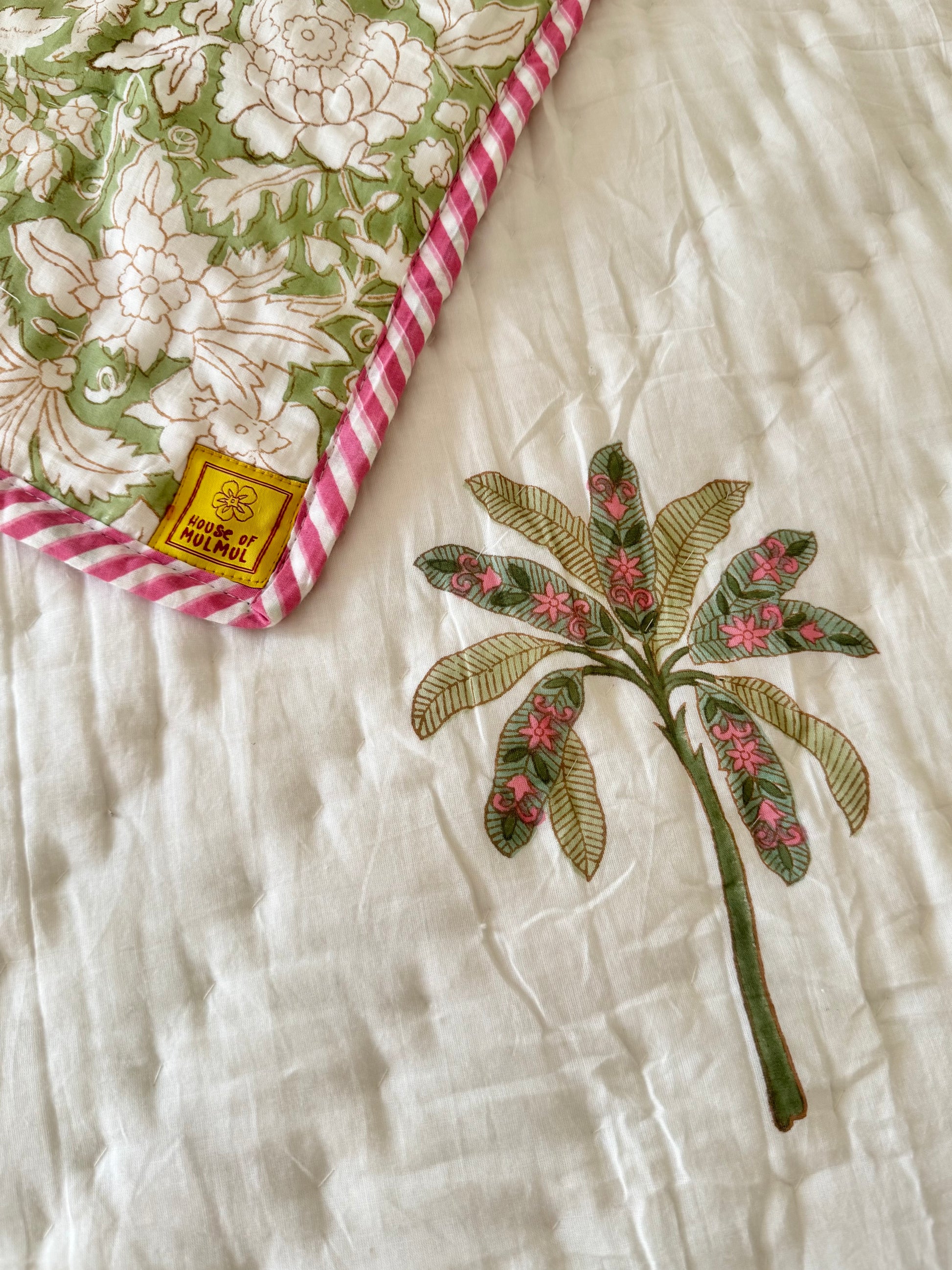 House of Mulmul - Blockprint Cotton Quilt Reversible. A close-up of a white quilted blanket featuring a green and pink embroidered palm tree motif, with a second matching quilt corner visible in the top-left showing a green floral pattern and pink striped border. A small yellow brand tag reads 'House of Mulmul'.