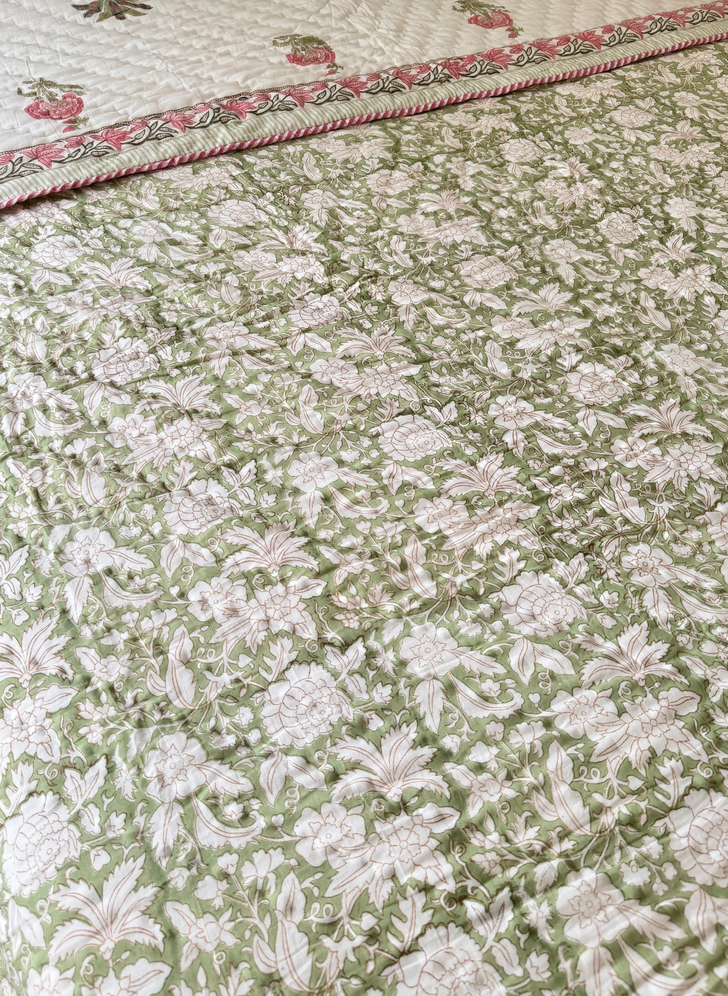 House of Mulmul - Blockprint Cotton Quilt Reversible. A close-up of a quilted cotton quilt featuring an all-over floral pattern in sage green and cream, with a decorative border showing pink and green floral details.