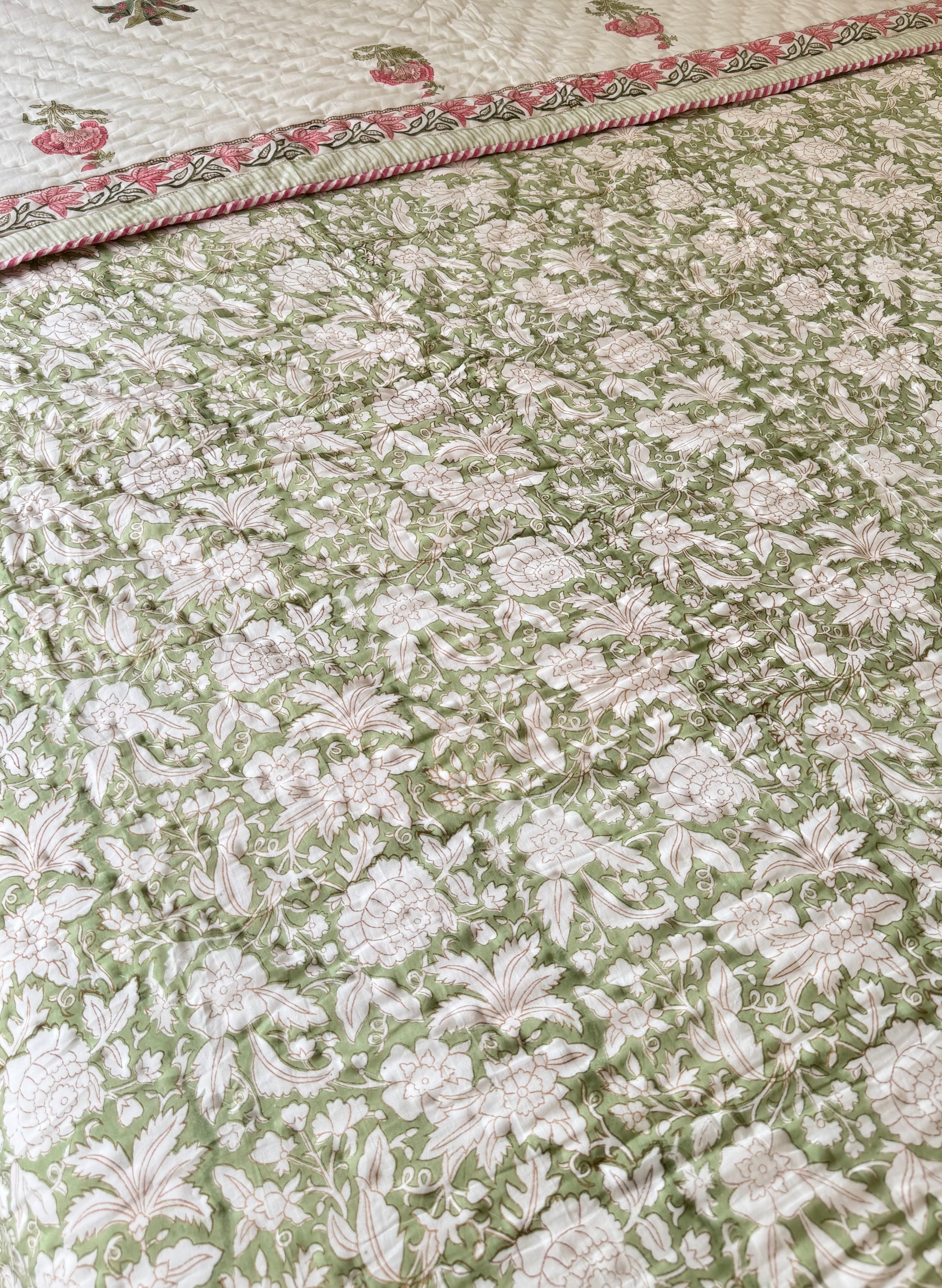 House of Mulmul - Blockprint Cotton Quilt Reversible. A close-up of a quilted cotton quilt featuring an all-over floral pattern in sage green and cream, with a decorative border showing pink and green floral details.