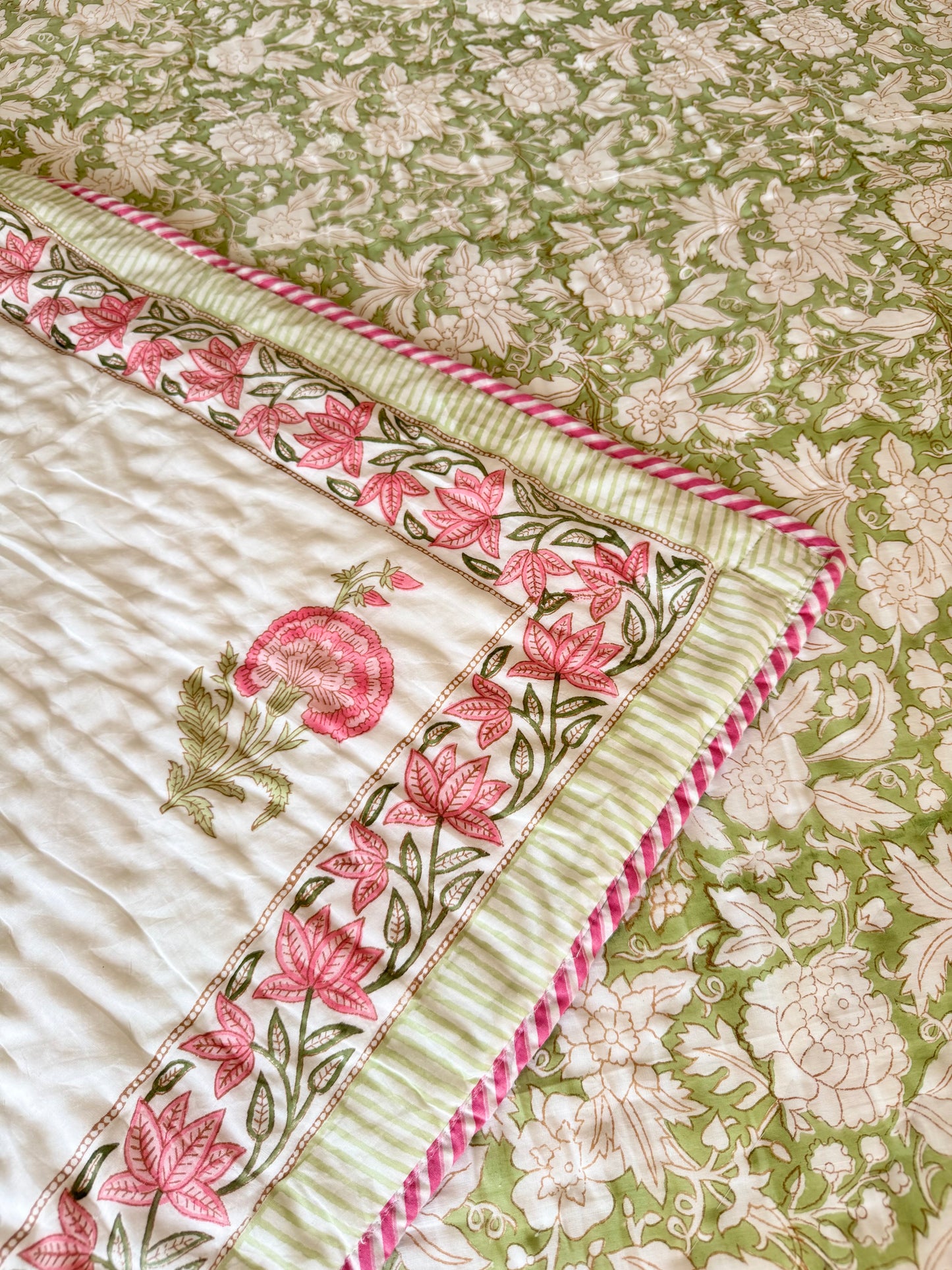 House of Mulmul - Blockprint Cotton Quilt Reversible. A close-up of a quilted cotton quilt with a floral print in sage green and cream, featuring a contrasting border with pink floral motifs and twisted pink-and-white edging.