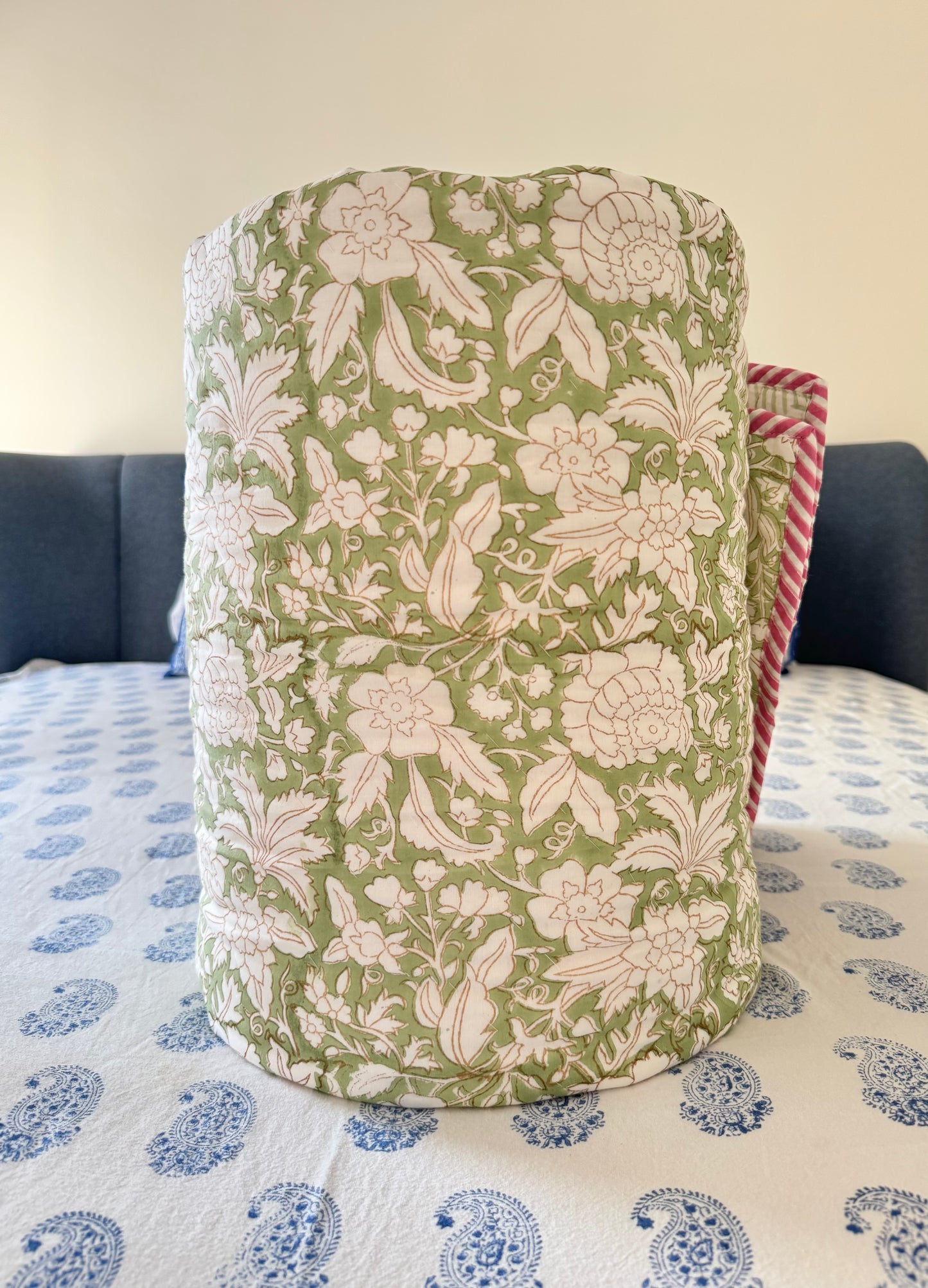 House of Mulmul - Blockprint Cotton Quilt Reversible. A cylindrical cotton quilt with a green and cream floral pattern, standing on a blue and white patterned bedspread in front of a dark headboard.