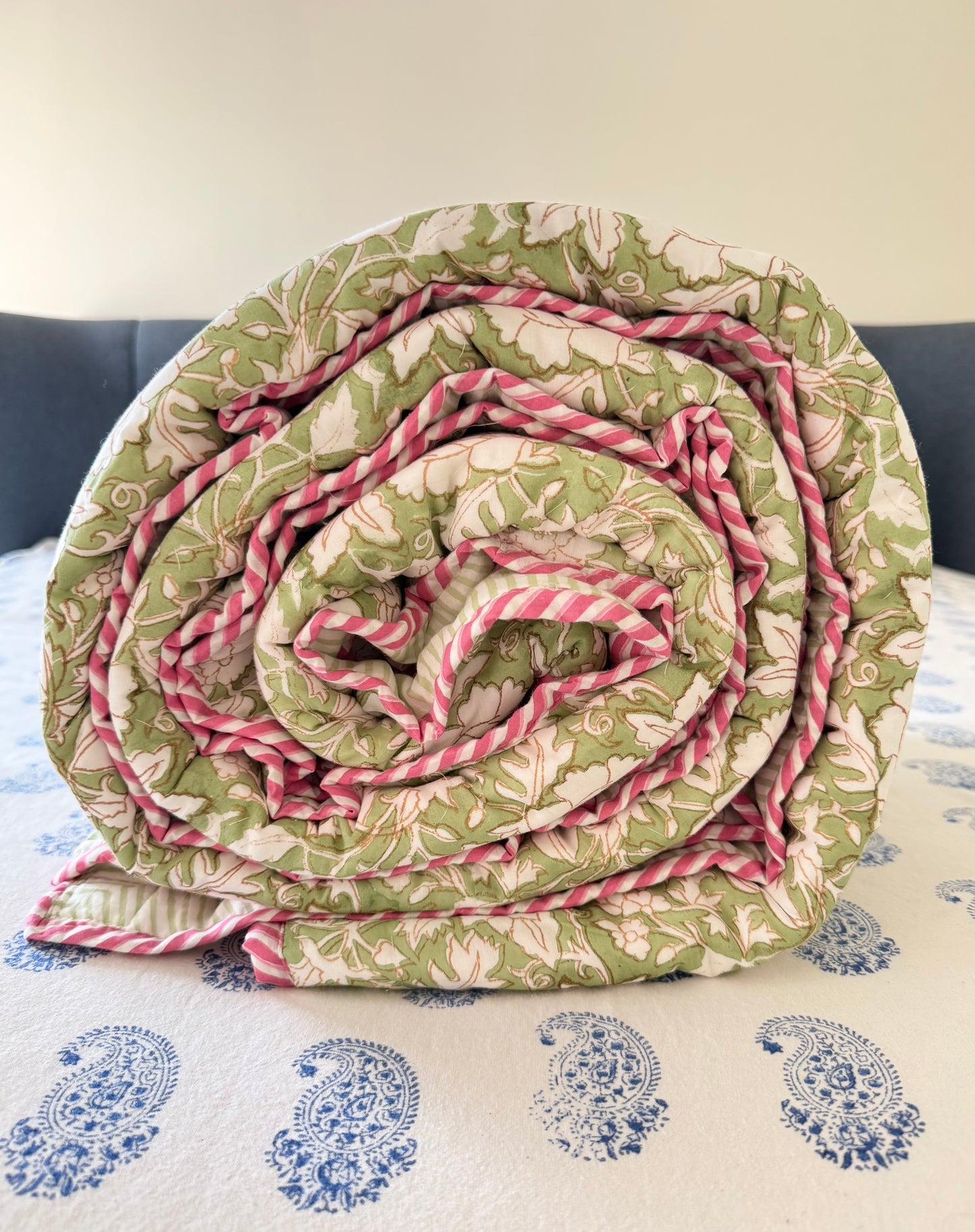 House of Mulmul - Blockprint Cotton Quilt Reversible. A rolled quilt or comforter with a green and beige botanical pattern and pink striped borders, displayed on a bed with a blue paisley-patterned sheet.