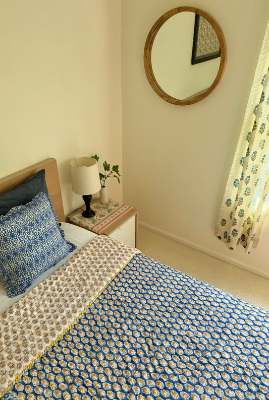 House of Mulmul - Blockprint Cotton Quilt Reversible. A neatly made bed in a bedroom with cream walls, featuring a blue and beige patterned block-print cotton bedspread, matching pillows, a bedside table with a lamp, and a round wooden wall mirror.