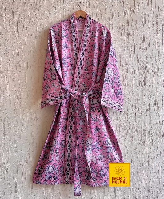 House of Mulmul - Blockprinted Cotton Bathrobe. A pink cotton robe with a black and purple floral block print pattern, hanging on a wooden hanger against a textured beige plaster wall. A yellow 'House of MUMUL' brand label is visible in the bottom right corner.