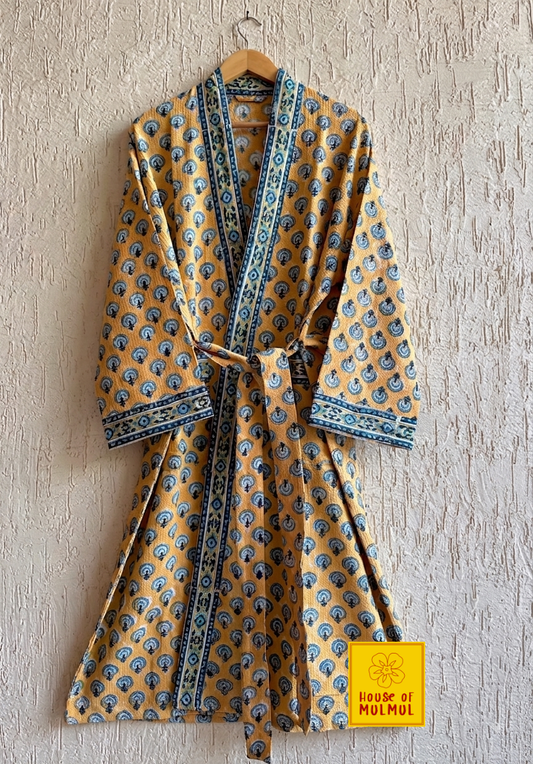 House of Mulmul - Blockprinted Cotton Bathrobe. A mustard yellow cotton robe with blue and black block print motifs and a blue border, hanging on a wooden hanger against a textured beige wall. A yellow 'House of MUMUL' brand label is visible in the bottom right corner.