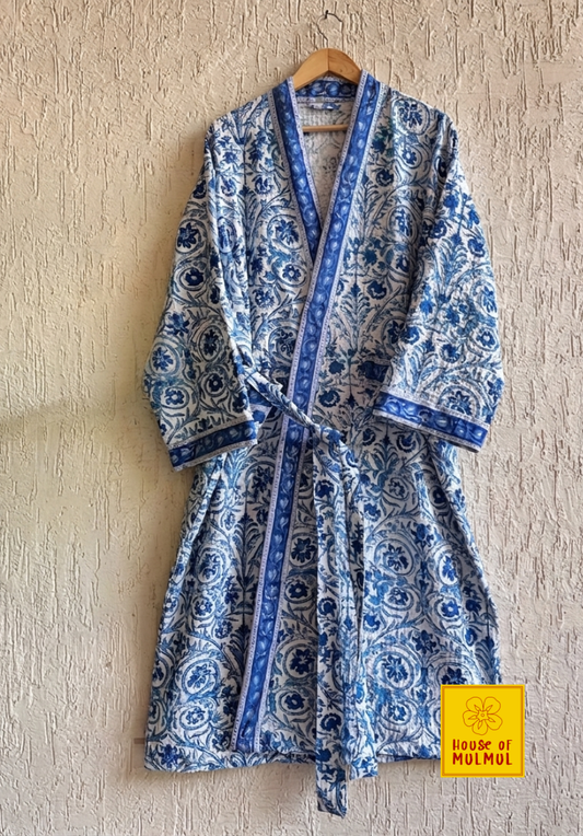 House of Mulmul - Blockprinted Cotton Bathrobe. A blue and white block-printed cotton robe with a matching tie belt, hanging on a wooden hanger against a textured beige wall. A yellow 'House of MULMUL' brand label is visible in the bottom right corner.
