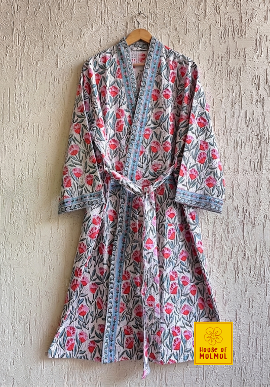 House of Mulmul - Blockprinted Cotton Bathrobe. A long-sleeved floral print kimono-style robe with a tie belt, hanging on a wooden hanger against a textured light gray wall. The robe features a white base with pink and red floral motifs and blue-gray leafy patterns, accented by a light blue patterned border.