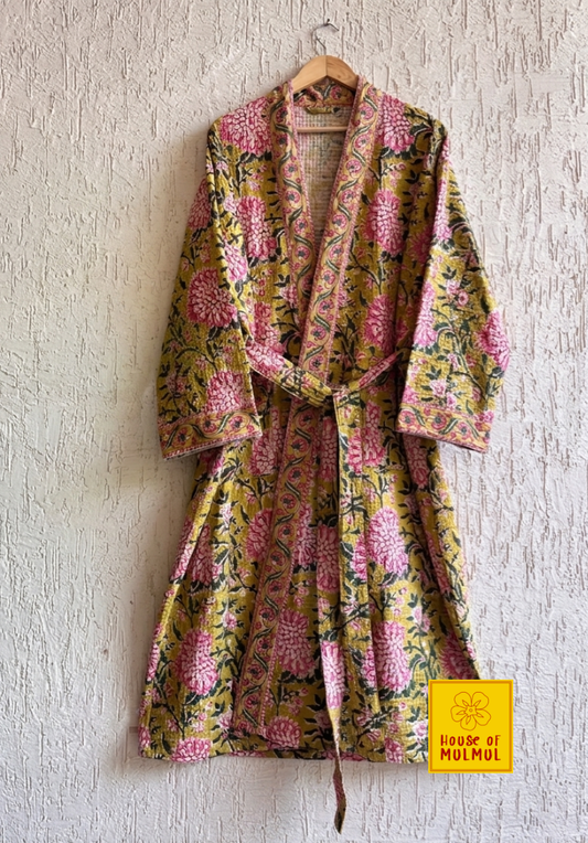 House of Mulmul - Blockprinted Cotton Bathrobe. A floral print cotton robe with pink chrysanthemums and green leaves on a mustard yellow background, hanging on a wooden hanger against a textured white wall. A yellow 'house of mulmul' logo is visible in the bottom right corner.