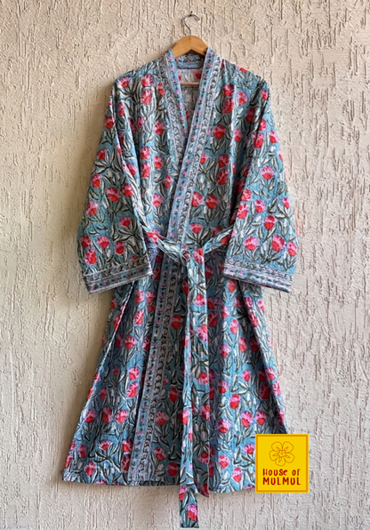 House of Mulmul - Blockprinted Cotton Bathrobe. A long-sleeved floral print robe hanging on a wooden hanger against a textured beige wall. The robe features a muted blue-gray base with pink and red flowers and green leaves, and has a matching tie belt.