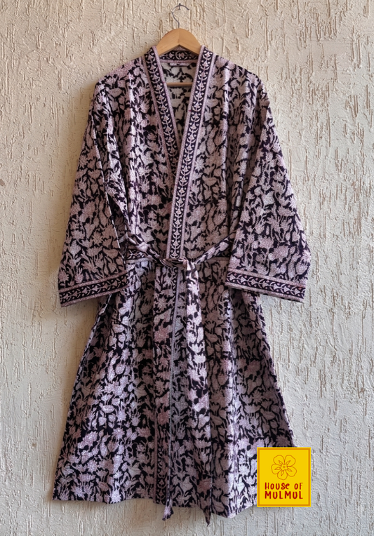 House of Mulmul - Blockprinted Cotton Bathrobe. A long-sleeved patterned robe with a matching tie belt hangs on a wooden hanger against a textured beige wall. The robe features a black and light lavender floral print with a dark patterned border.