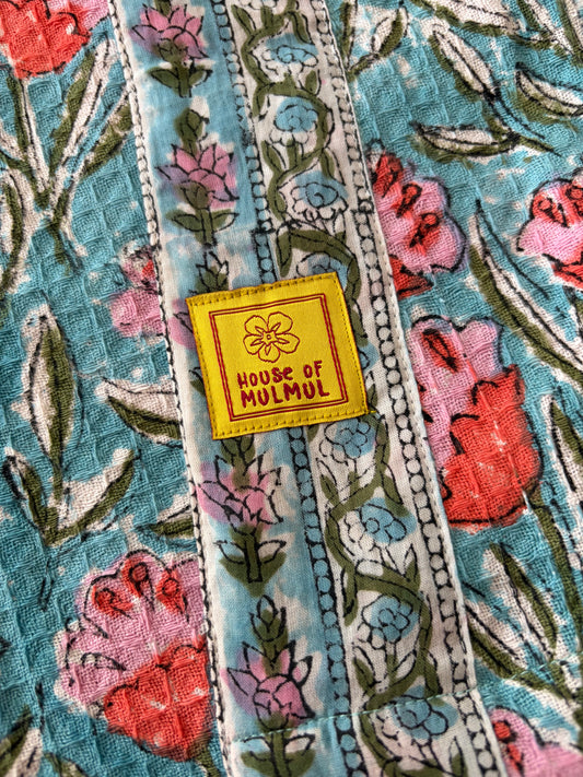 House of Mulmul - Blockprinted Cotton Bathrobe. A close-up of a colorful floral bathrobe with vertical patterned bands and a yellow square patch reading 'HOUSE OF MULMUL'.