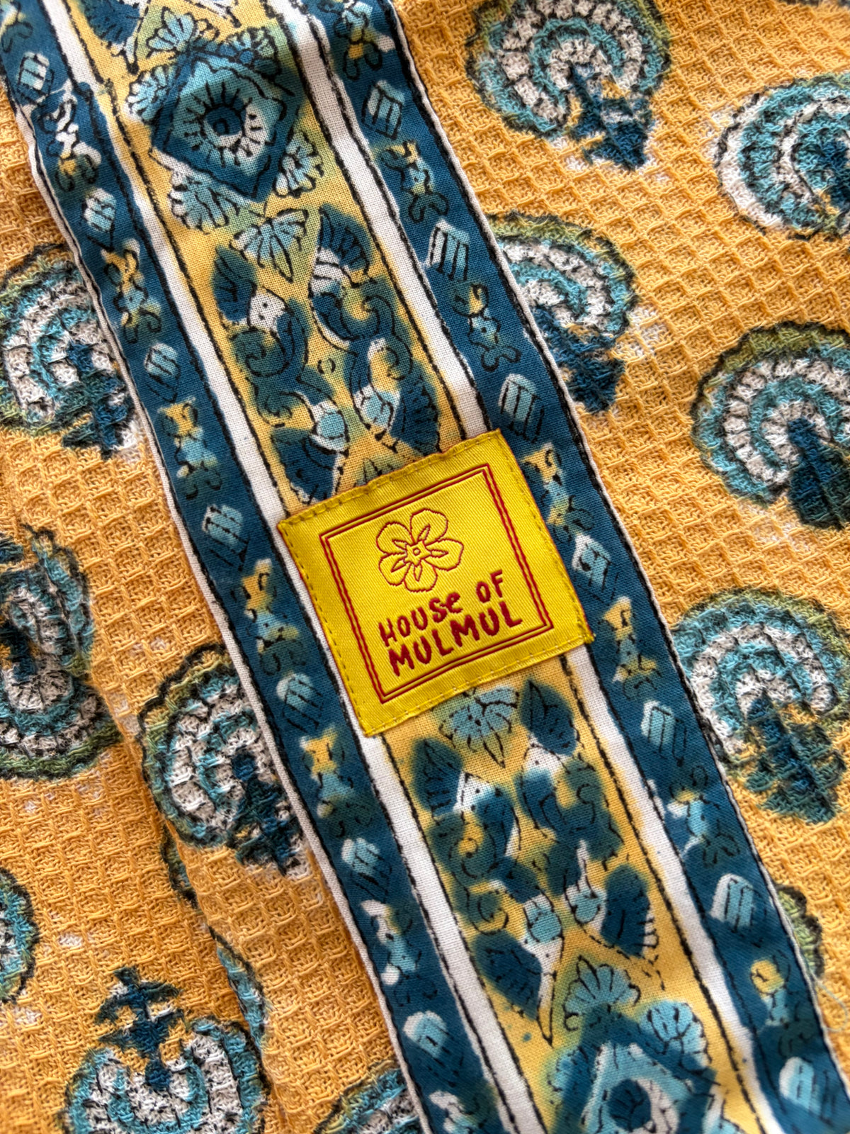 House of Mulmul - Blockprinted Cotton Bathrobe. A close-up of a patterned bathrobe featuring a yellow waffle-weave base with teal and blue paisley motifs, bordered by striped bands, and a central yellow patch reading 'HOUSE OF MULMUL'.