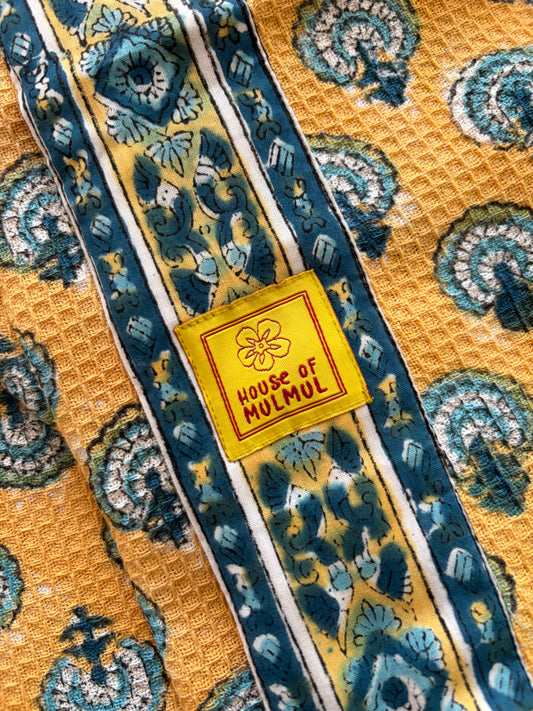 House of Mulmul - Blockprinted Cotton Bathrobe. A close-up of a patterned bathrobe featuring a yellow waffle-weave base with teal and blue paisley motifs, bordered by striped bands, and a central yellow patch reading 'HOUSE OF MULMUL'.