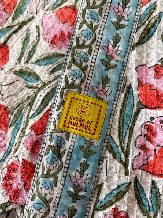 House of Mulmul - Blockprinted Cotton Bathrobe. A close-up of a colorful handblock printed bathrobe with a yellow square label reading 'HOUSE OF MULMUL'. The textile features floral and vine patterns in pink, red, light blue, and green on a cream background.
