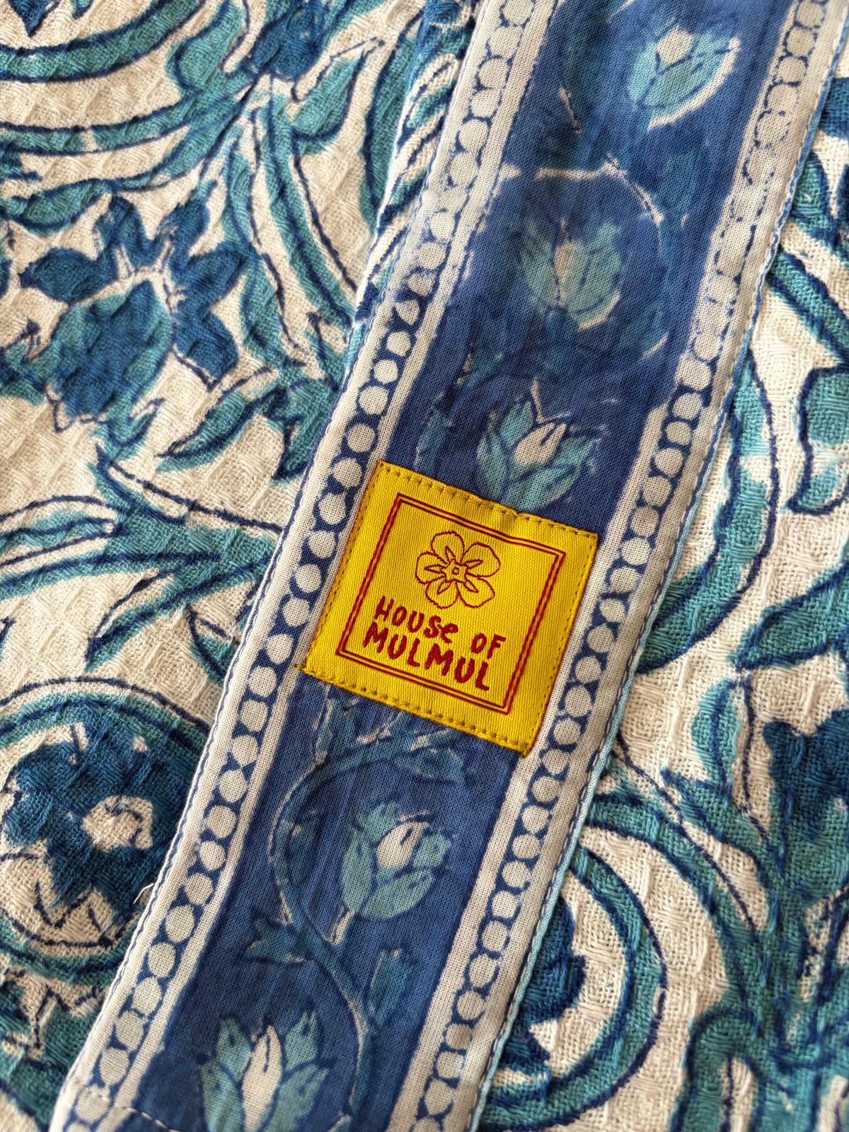 House of Mulmul - Blockprinted Cotton Bathrobe. A close-up of a blue and cream block-printed bathrobe with a yellow woven label reading 'HOUSE OF MUMUL'.