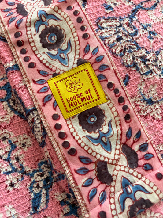 House of Mulmul - Blockprinted Cotton BathrobeA close-up of a pink patterned bathrobe with ornate floral and paisley motifs in blue, cream, and dark brown, featuring a yellow sewn label that reads 'HOUSE OF MUMUL'.