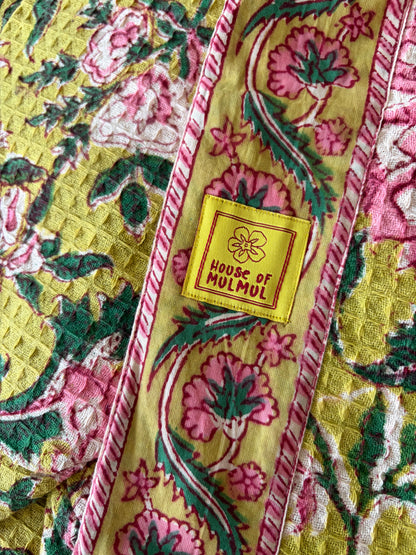 House of Mulmul - Blockprinted Cotton Bathrobe. A close-up of a quilted bathrobe, likely a shawl or blanket, featuring a yellow base with pink and green floral motifs and a central yellow label that reads 'HOUSE OF MULMUL'.