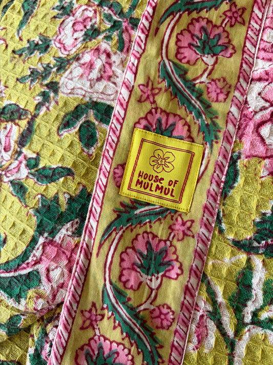 House of Mulmul - Blockprinted Cotton Bathrobe. A close-up of a quilted bathrobe, likely a shawl or blanket, featuring a yellow base with pink and green floral motifs and a central yellow label that reads 'HOUSE OF MULMUL'.