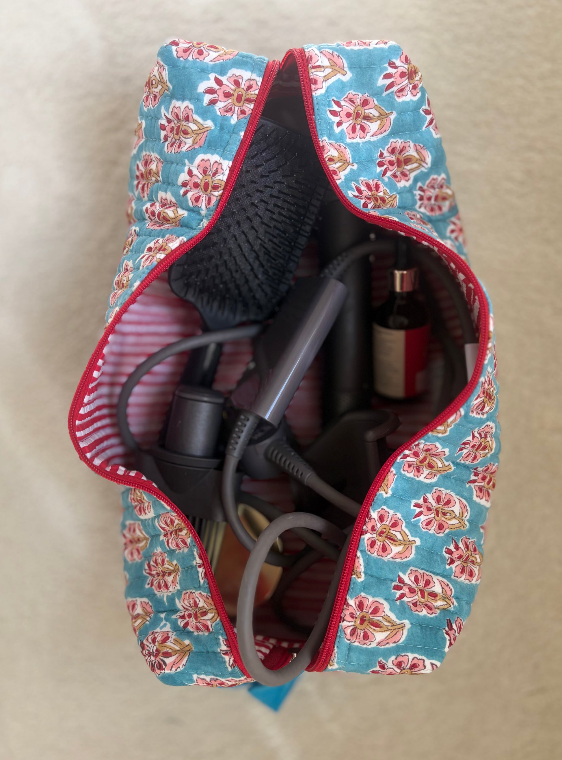 House of Mulmul - Blockprinted Cotton Hair Tool Bag. Floral-patterned  Dyson hair dryer straightener kit hair tool bag containing a hair dryer, bottle, and other items on a beige surface.