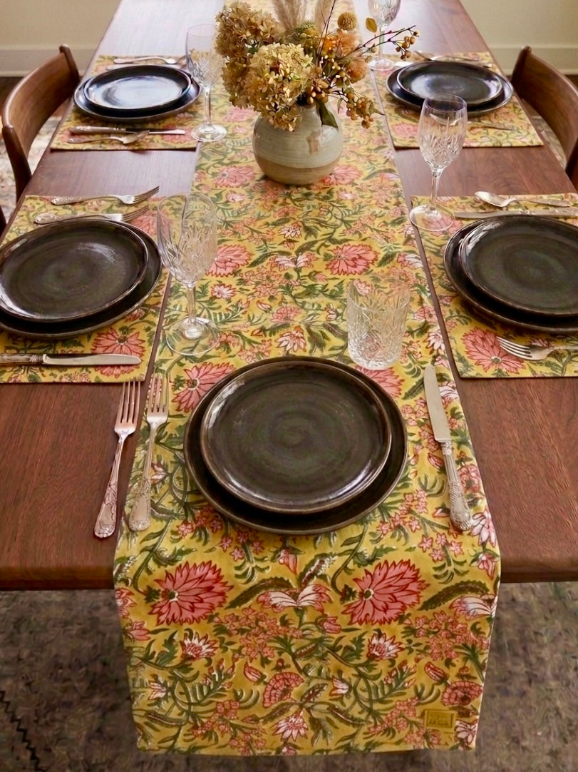 House of Mulmul - Blockprinted Cotton Reversible Table Runner & Mats set. Dining table set with floral table runner, black plates, and silverware.