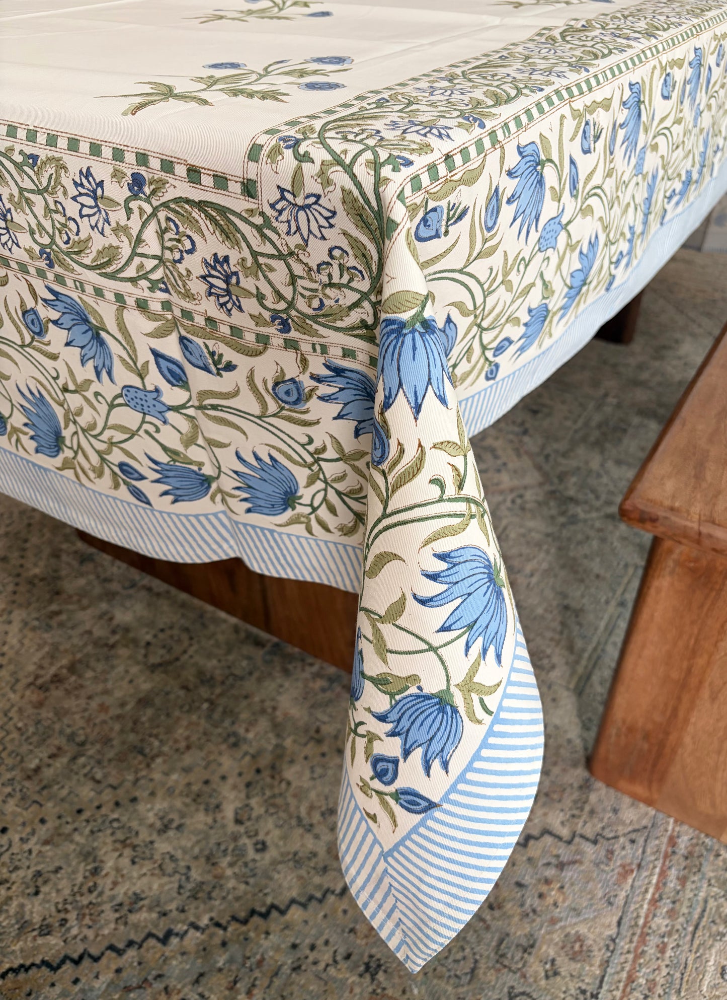 House of Mulmul - Blockprinted Cotton Table Cloth. A cream-colored tablecloth with a blue and green botanical floral pattern and striped borders, draped over a wooden table on a patterned rug.