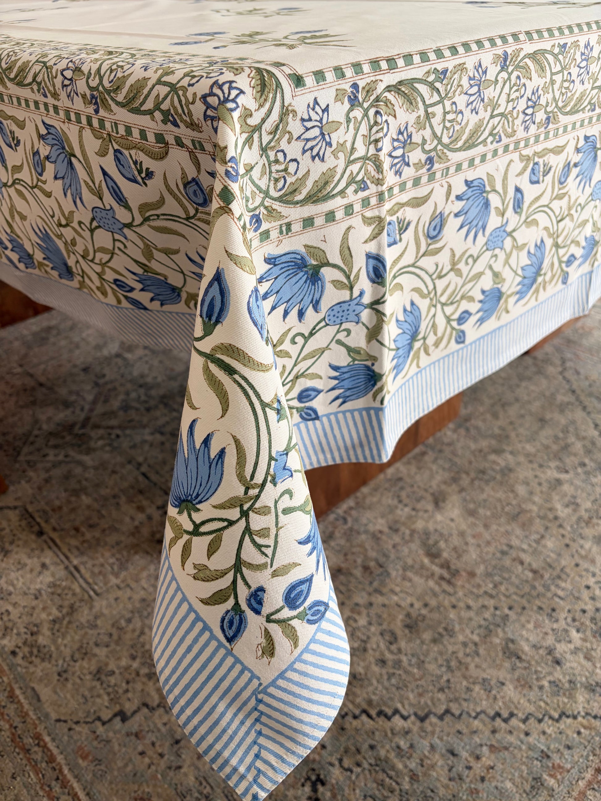 House of Mulmul - Blockprinted Cotton Table Cloth. A cream-colored tablecloth with a blue and green floral vine pattern and striped borders, draped over a table on a patterned rug.