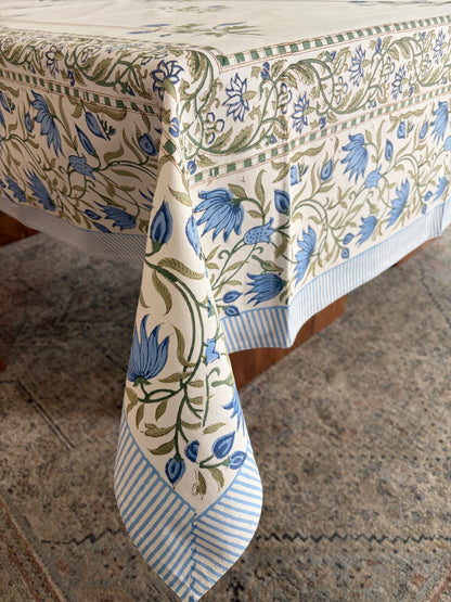 House of Mulmul - Blockprinted Cotton Table Cloth. A cream-colored tablecloth with a blue and green floral vine pattern and striped borders, draped over a table on a patterned rug.