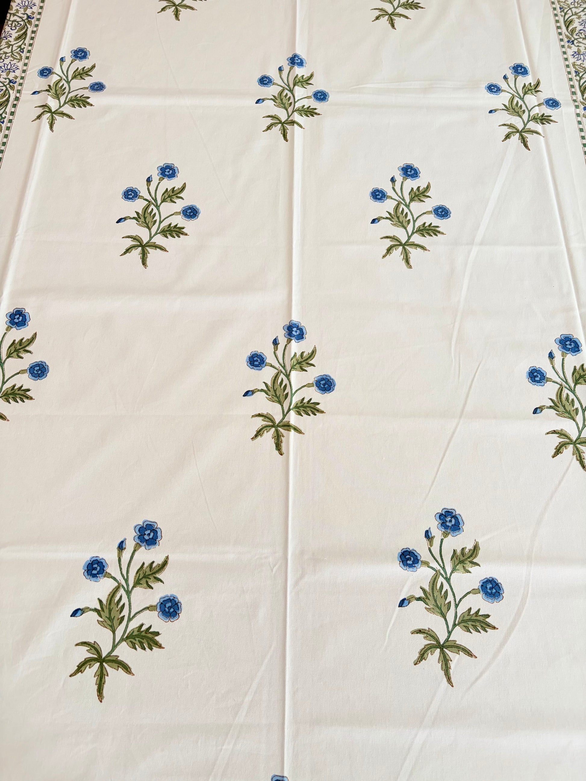 House of Mulmul - Blockprinted Cotton Table Cloth. An off-white cotton tablecloth featuring a repeating pattern of small blue flowers with green stems and leaves, laid flat.