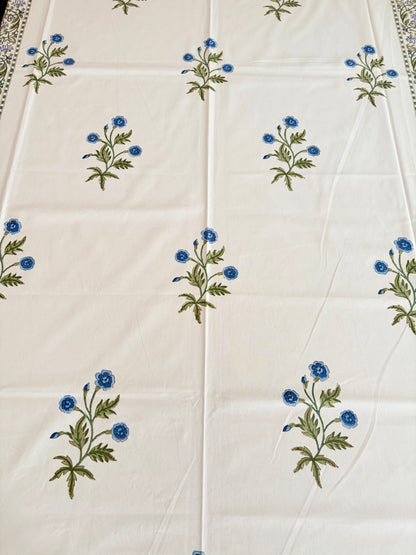 House of Mulmul - Blockprinted Cotton Table Cloth. An off-white cotton tablecloth featuring a repeating pattern of small blue flowers with green stems and leaves, laid flat.