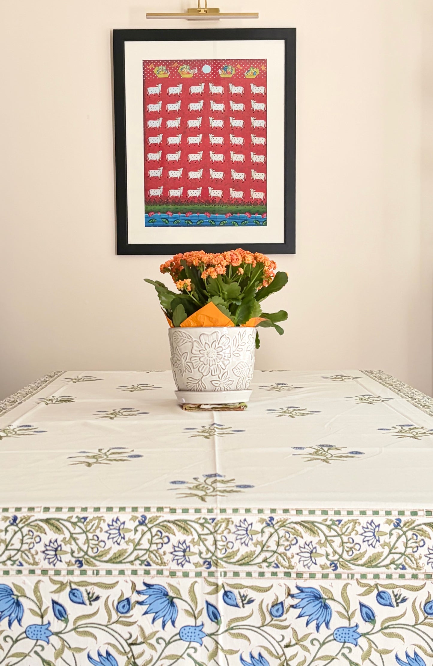 House of Mulmul - Blockprinted Cotton Table Cloth. A framed art print featuring a red and blue pattern of sheep hangs on a beige wall above a table covered with a matching blue and green floral tablecloth. A white patterned pot with orange flowers sits on the table.