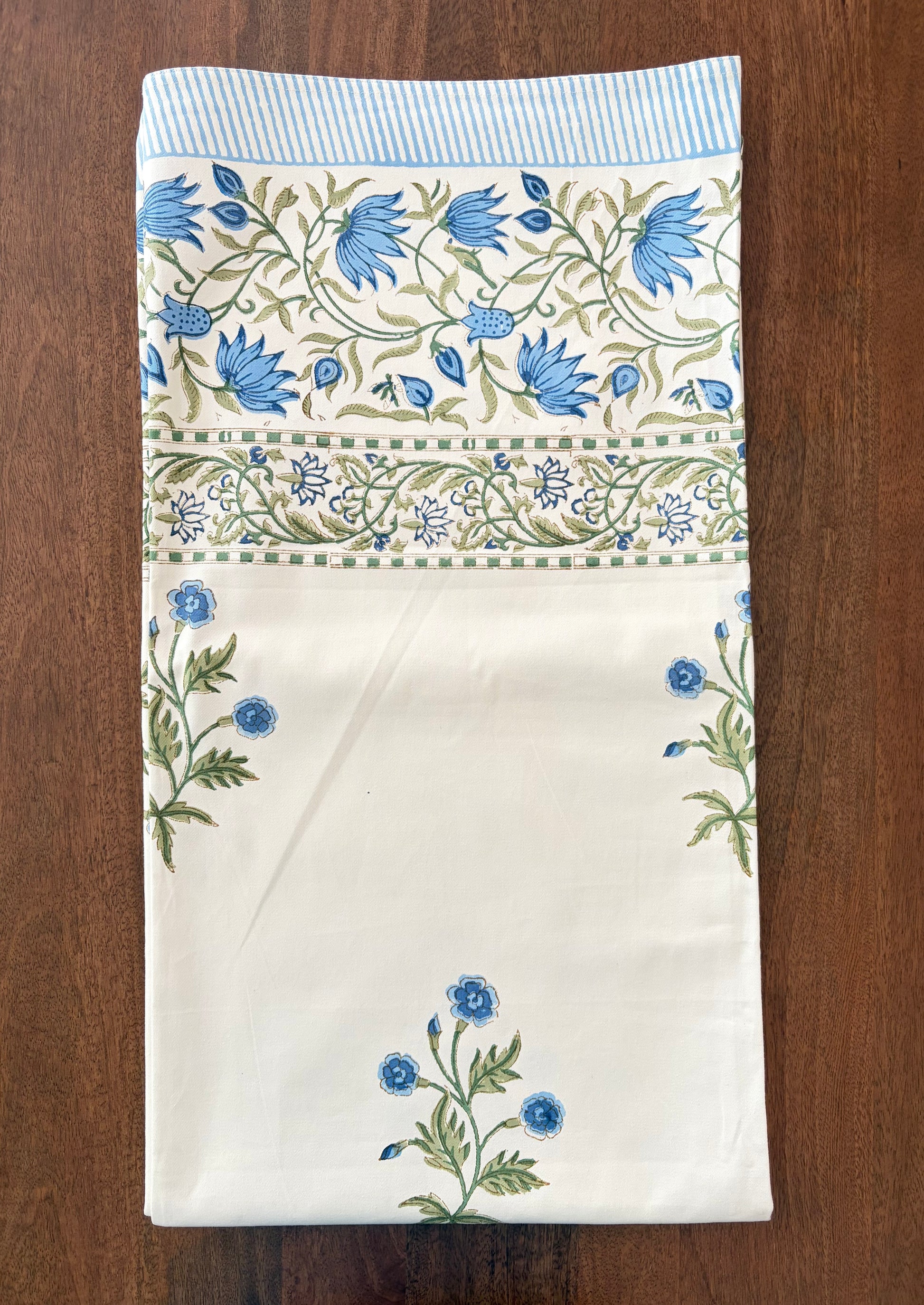 House of Mulmul - Blockprinted Cotton Table Cloth. A folded cream-colored cotton tablecloth with blue floral patterns and green leaves, laid flat on a dark brown wooden surface.