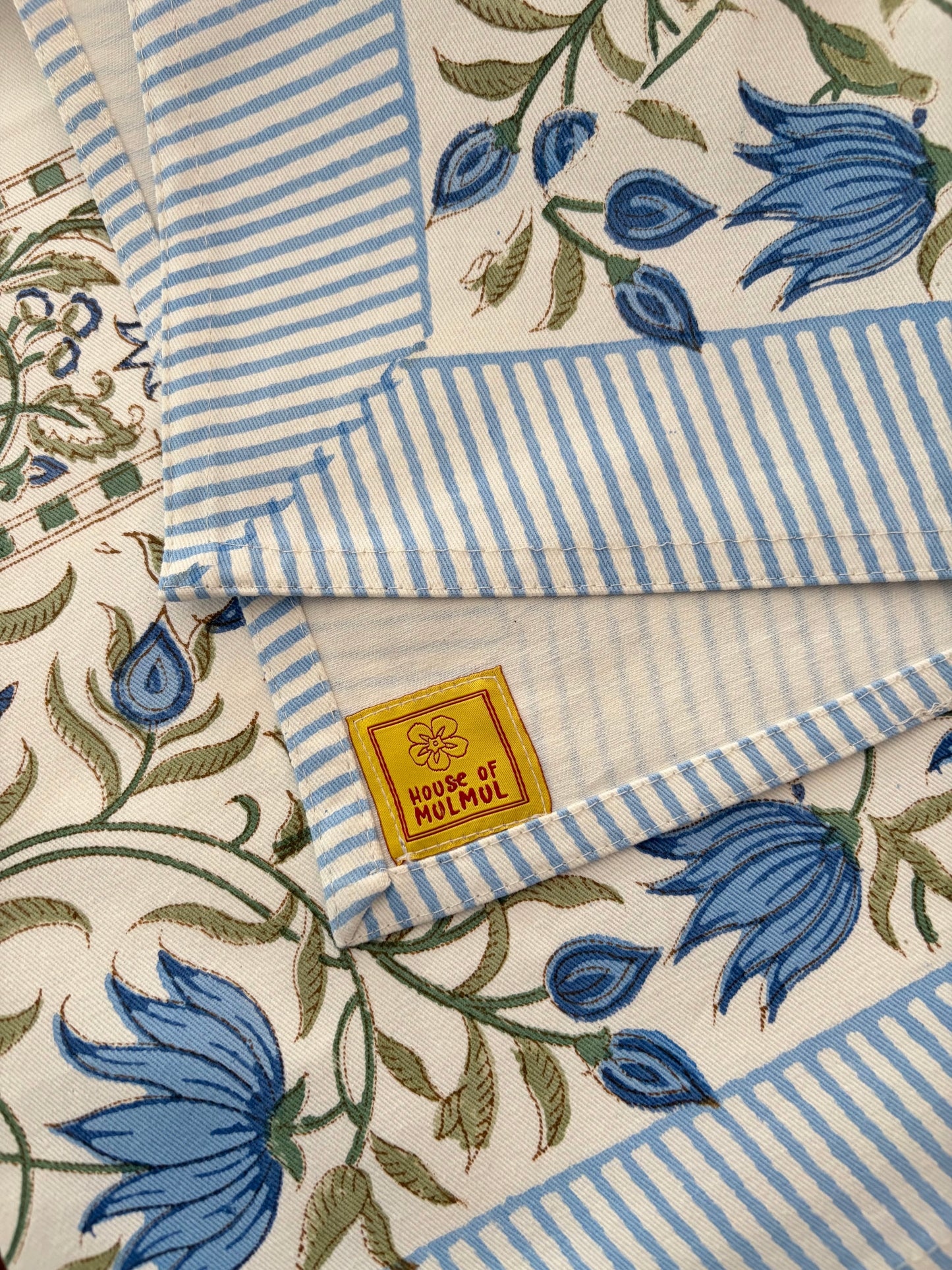 House of Mulmul - Blockprinted Cotton Table Cloth. A folded textile product showing a cream base with blue floral motifs, blue-and-white striped borders, and a small yellow brand label reading 'HOUSE OF MULMUL'.
