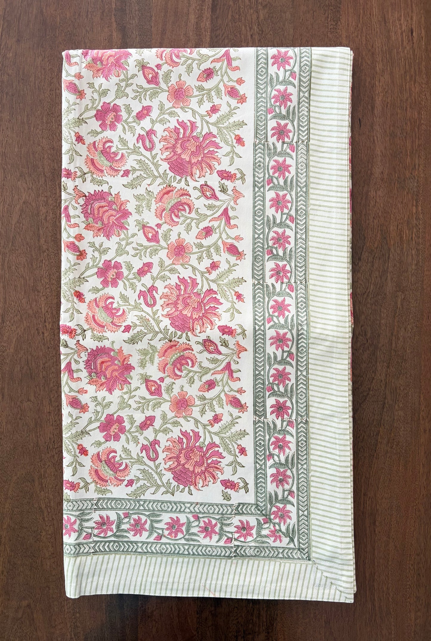 House of Mulmul - Blockprinted Cotton Table Cloth. A rectangular cream-colored textile, likely a tablecloth or napkin, featuring a vibrant pink and green floral pattern with a striped border, laid flat on a dark wood surface.